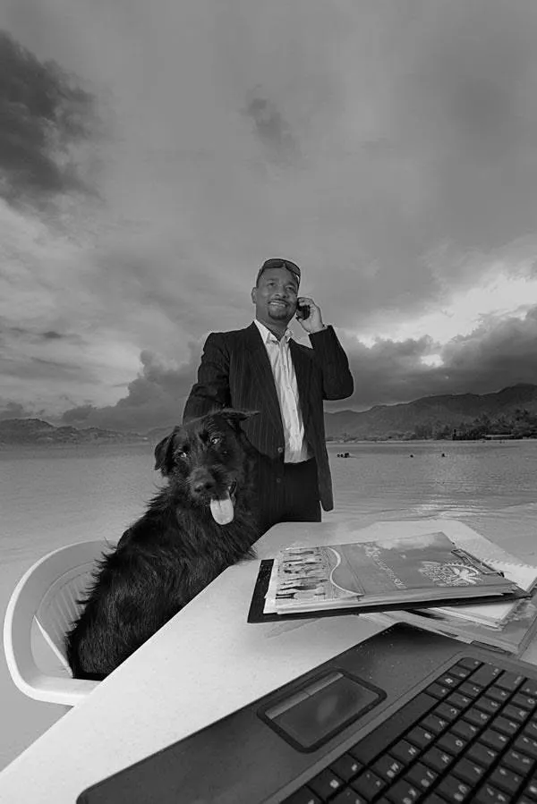 Black-and-white portrait of a Sunquest broker in a suit, smiling on the phone at a beachside desk, a black dog sitting beside him